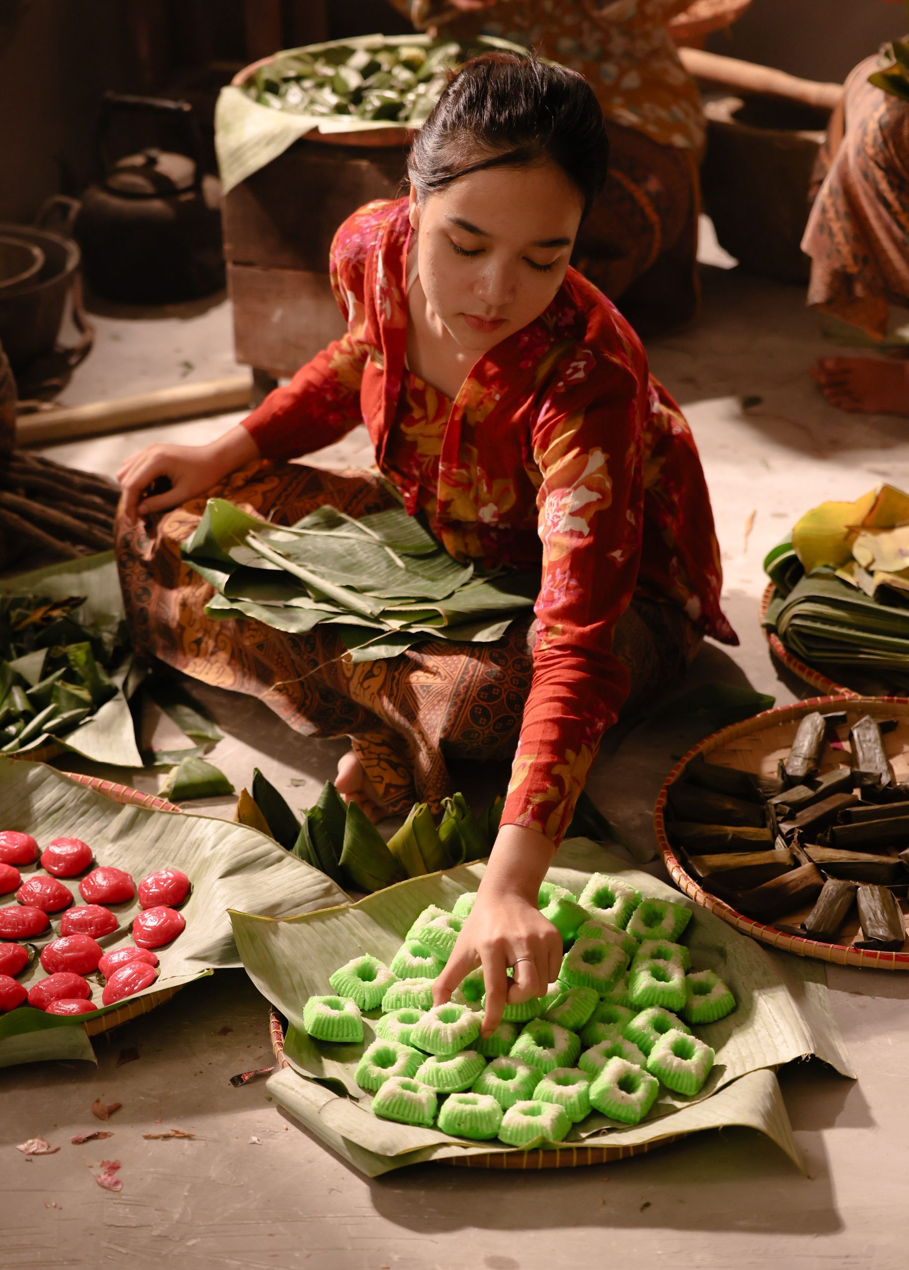 Asian woman arranging colorful traditional sweets in a market setting using banana leaves.