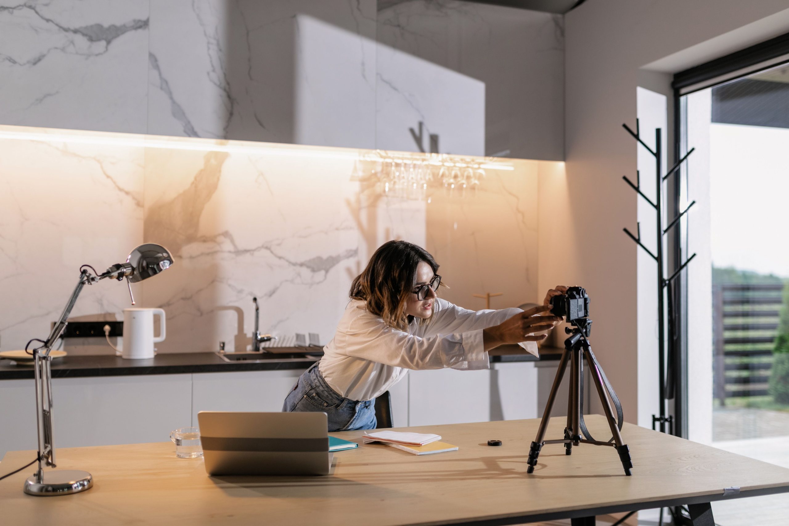 Young woman adjusting a camera on a tripod in a stylish kitchen, with natural lighting. Perfect for creative or lifestyle themes.